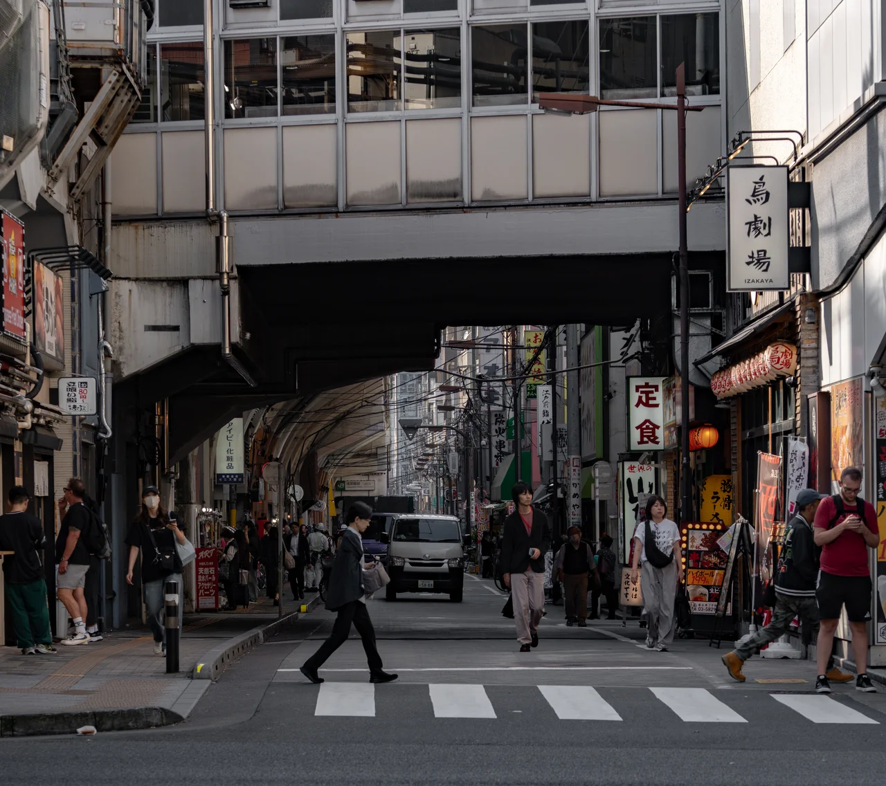 Un passant traverse un passage piéton sous une voie ferrée aérienne, entre des enseignes d'izakaya.