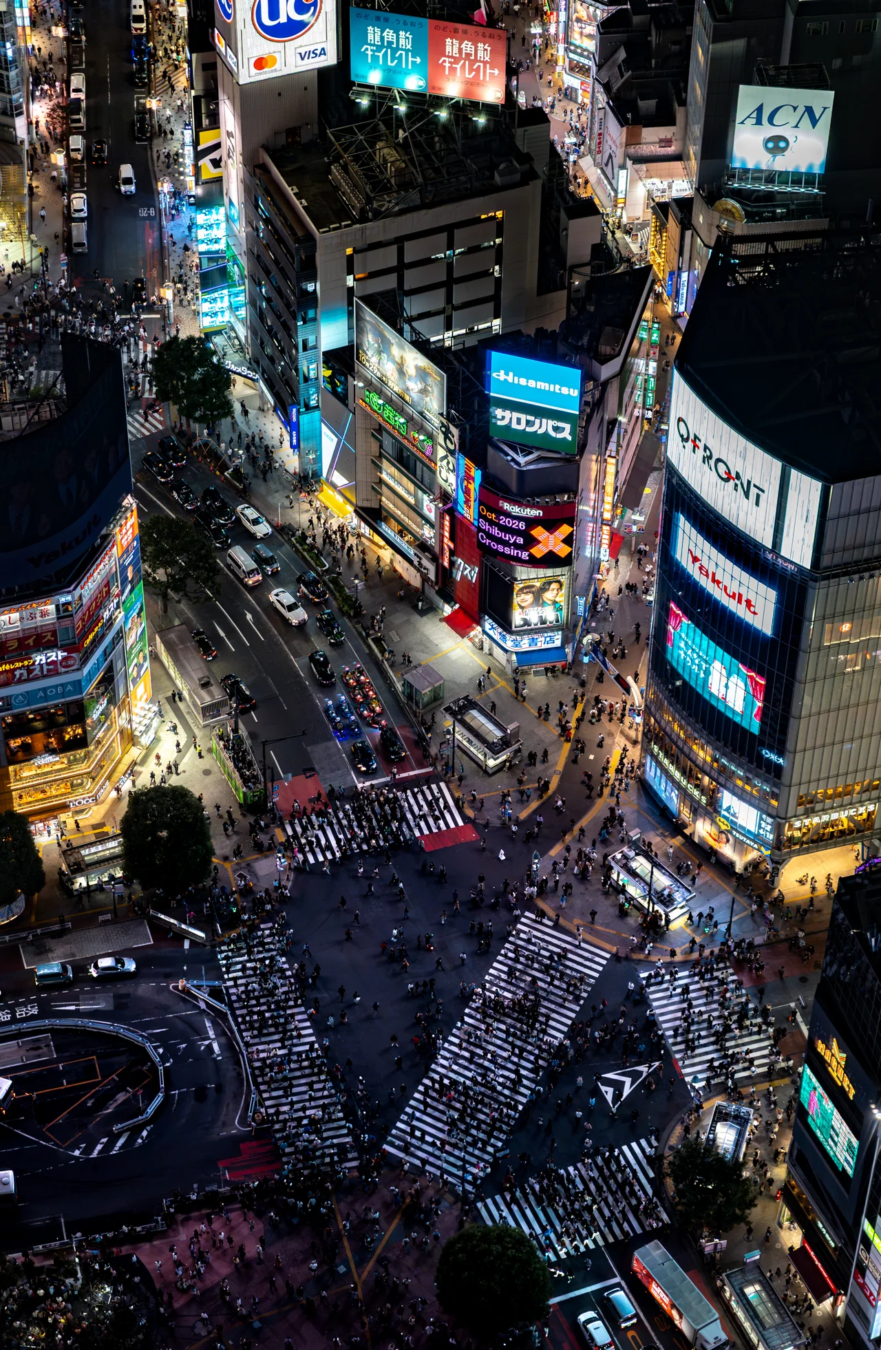 Vue plongeante de nuit sur le carrefour de Shibuya, ses passages piétons et ses écrans.